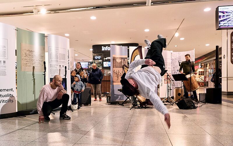 Im Vordergrund springt ein Tänzer durch die Luft, im Hintergrund stehen drei Musiker und eine Ausstellung.